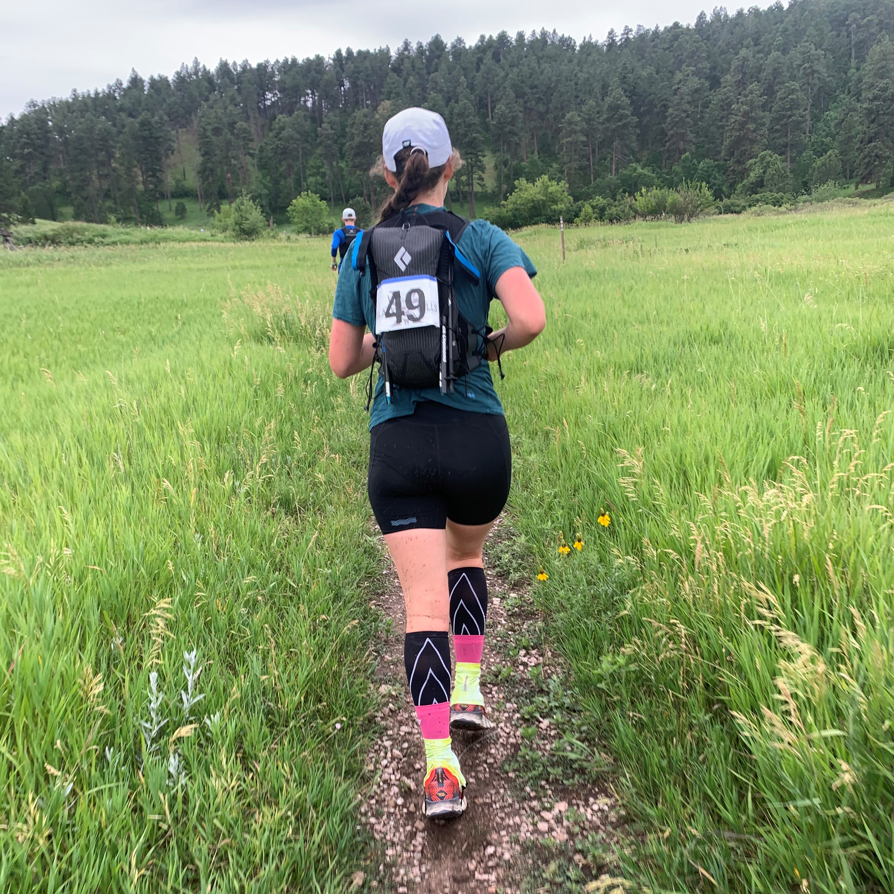 woman running away from camera on grassy trail, mountains in background