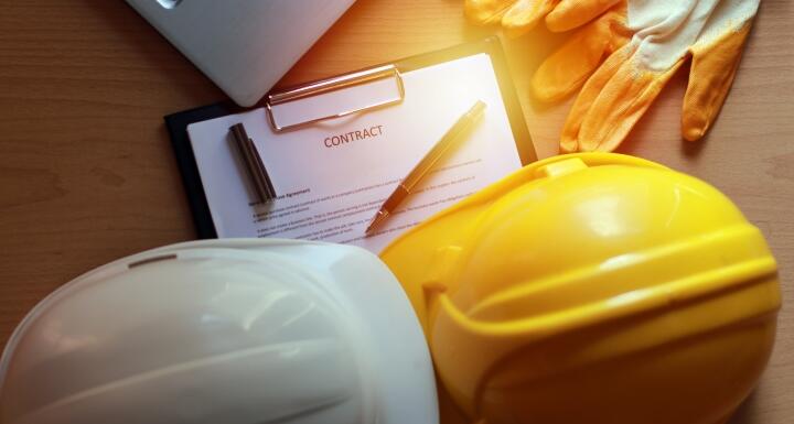 2 hard hats, a contract, gloves, and laptop on desk