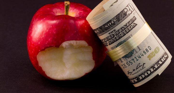 American currency leans against a red apple with a bite taken out of it