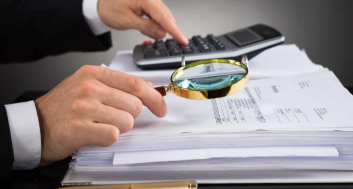 Close-up Of Businessperson Hands Checking Invoice With Magnifying Glass At Desk