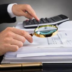 Close-up Of Businessperson Hands Checking Invoice With Magnifying Glass At Desk