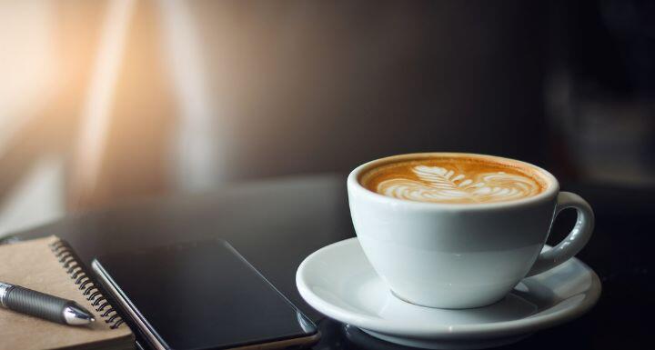 Closeup latte art coffee in white cup with smartphone and notebook on black table