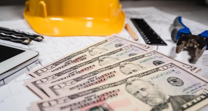 Yellow construction hat sitting on table with a layer of twenty dollar bills and a ruler and pencil