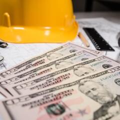 Yellow construction hat sitting on table with a layer of twenty dollar bills and a ruler and pencil