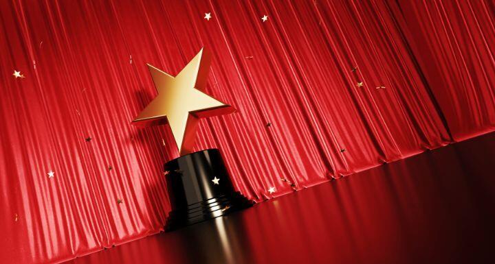 Falling Onto A Gold Star Shaped Award Sitting Before Red Stage Curtain 