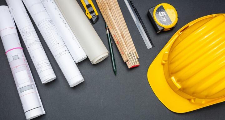 Yellow hard hat blue prints tape measure pen and rulers laying on a black surface