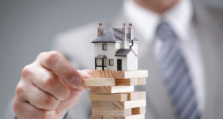 Hand of a man in a suit removing a block from a jenga game with a house sitting on top