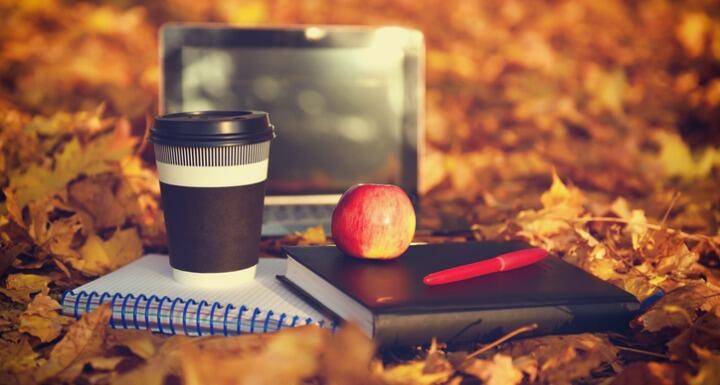 Laptop, apple, coffee, notepads, and pens surrounded by fall foliage