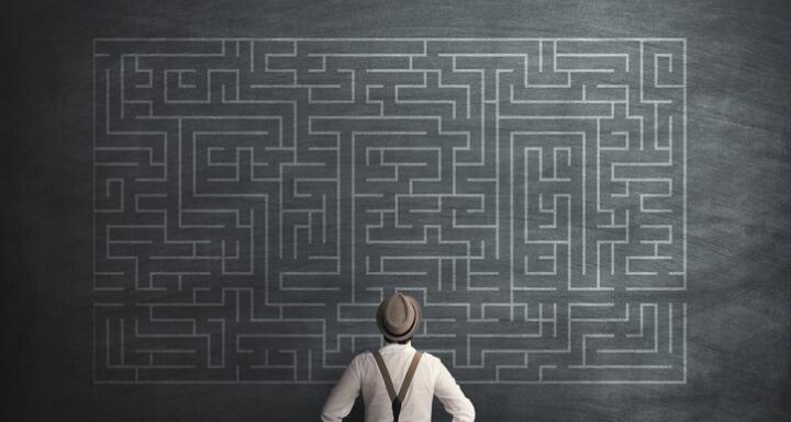 Man in hat and suspenders standing in front of a chalk board with an intricate maze written on it