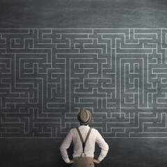Man in hat and suspenders standing in front of a chalk board with an intricate maze written on it