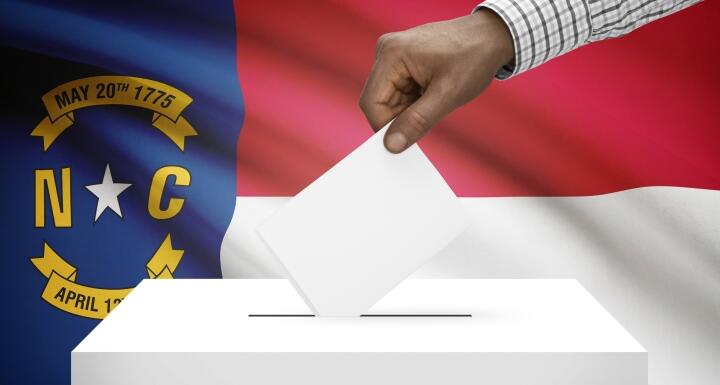 Person's hand dropping a white ballot into a white ballot box with the North Carolina State flag set as the back drop