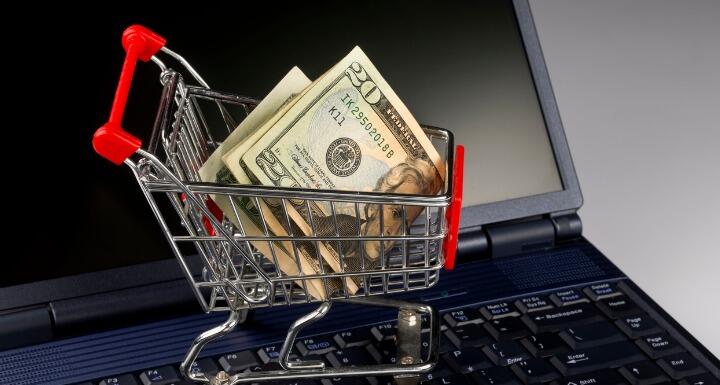 Tiny metal shopping cart sitting on the keyboard of a laptop with folded twenty dollars in it