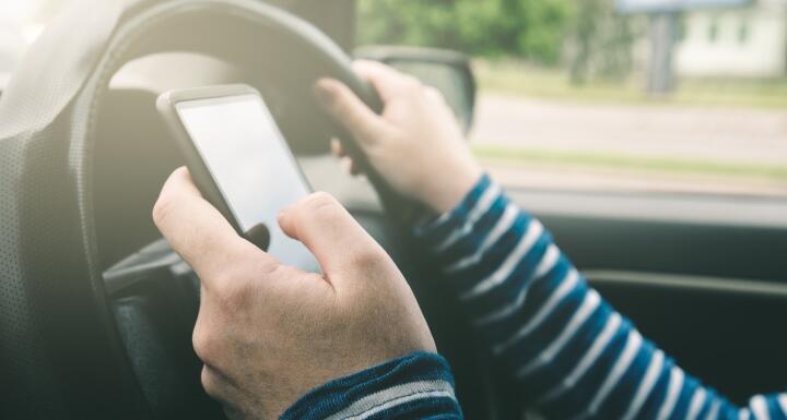 Person texting on cell phone in left hand and holding car steering wheel with right hand