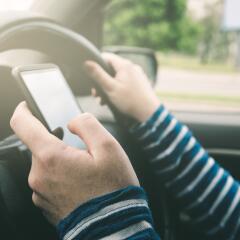 Person texting on cell phone in left hand and holding car steering wheel with right hand