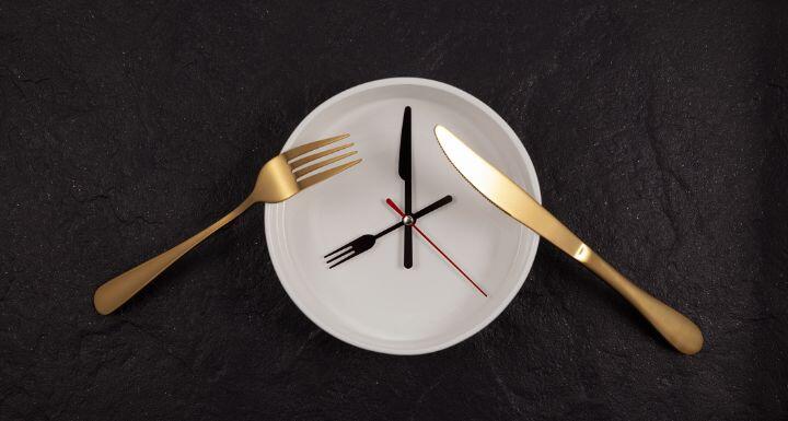 White deep plate, knife and fork. Clock on a dark textured stone background with copy space