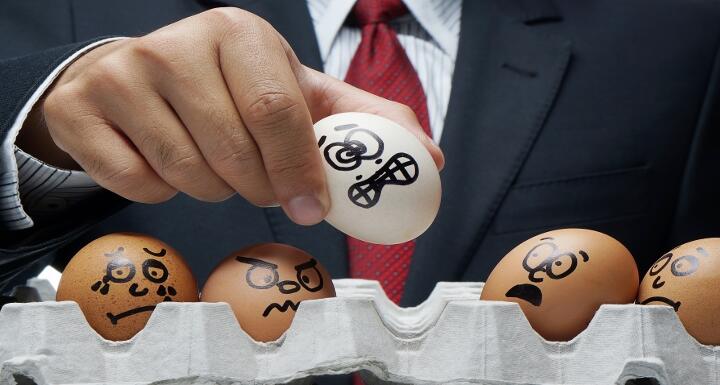 Man in business suit holding egg with an unhappy face drawn on it as other eggs remain in container