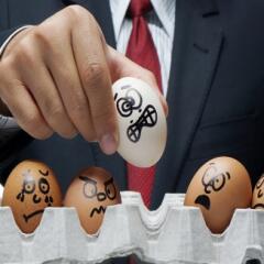 Man in business suit holding egg with an unhappy face drawn on it as other eggs remain in container