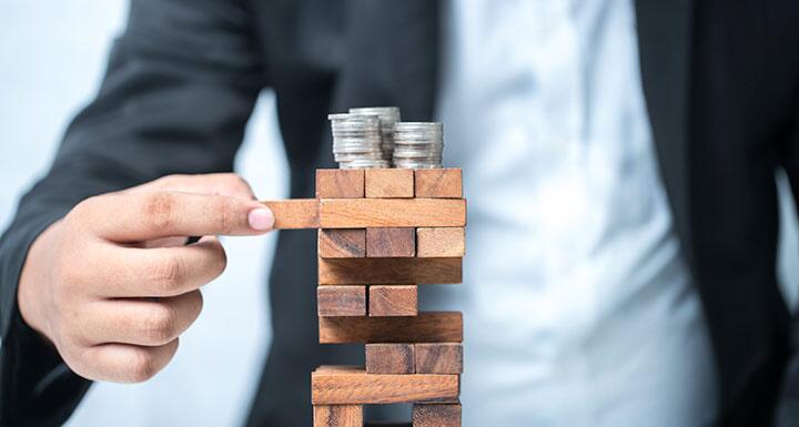 Stack of Jenga blocks with three stacks of coins on top and a man in a suit pulling one of the blocks from near the top of the stack