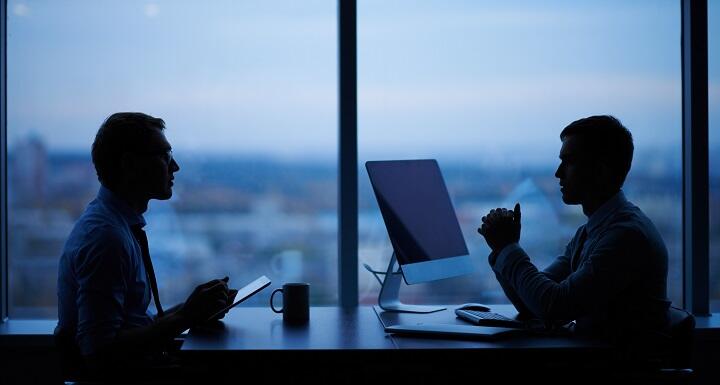 Two shadowy figures talking in front of large office windows